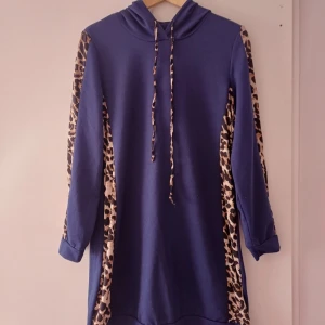 Blå klänning med leopardmönster -  Använd men ser ut som ny. En blå klänning med huva och leopardmönstrade detaljer på ärmarna. Klänningen har långa ärmar och en avslappnad passform.