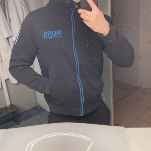 Hugo Boss Hoodie, svart och blå - Svart hoodie från Hugo Boss med blåa detaljer. Hoodien är i storlek S och är i bra skick. Köpt i Mall of scandinavia på Hugo Boss. 