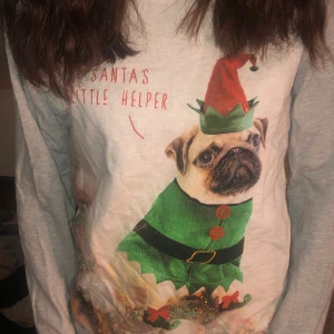 En vit tröja med en hund och står en text - Det är en vit tröja med röd text där de står santas littel helper och texten kommer fåren en hund med Nisse kostym