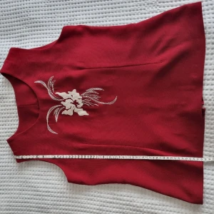 Vintage röd väst - Höj din outfit med denna vintage röda väst som skriker 80-tals sofistikation! Den högkvalitativa tyget och den kamferade midjan gör den till ett framstående plagg. Har en vit blommig brodyr på bröstet som lägger till en toucjmh av tidlös glamour. 