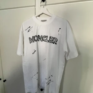 Vit Moncler t-shirt med graffiti-tryck - Säljer en vit t-shirt från Moncler med coolt svart graffiti-inspirerat tryck både fram och bak. T-shirten har rund hals, korta ärmar och är gjord i mjuk bomull. Perfekt för dig som gillar streetstyle och vill sticka ut med ett unikt plagg.
