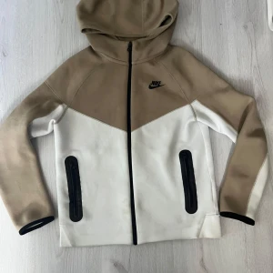  beige nike tech barn 147-158 - Snygg vindjacka från Nike i beige och vitt med svart logga på bröstet. Jackan har huva, hel dragkedja och två svarta fickor med dragkedja framtill. Perfekt för dig som gillar sportig stil och vill ha en jacka som sticker ut med sin coola färgblock-design.