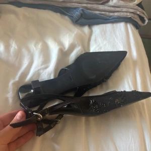 Svarta spetsiga pumps från MNG strl 37 - Svarta pumps från MNG med spetsig tå och låg klack. Skorna har snygga paljett- och pärldetaljer på ovandelen samt smal rem med spänne runt hälen. Materialet är syntet och textil, vilket ger en cool och elegant look.