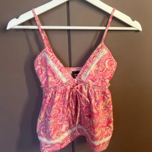 Rosa mönstrad linnetopp Gina Tricot - Söt linnetopp från Gina Tricot i rosa och beige med ett unikt mönster. Toppen har smala axelband, v-ringning och dekorativa spetsdetaljer samt knyt framtill. Luftig och lätt i bomullsmaterial.