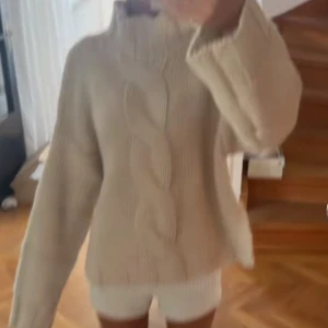 Beige stickad polotröja med flätmönster - Supermysig beige stickad polotröja med grovt flätmönster framtill. Tröjan har en hög krage och långa ärmar, perfekt för kyliga dagar. Den är loose i passformen och riktigt skön att bära till jeans eller leggings.