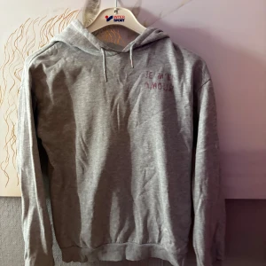 Grå hoodie med tryck från Gina Tricot - Grå hoodie från Gina Tricot med texten 'Je m'en amour' tryckt i rött och svart på bröstet och större på ryggen. Mjuk bomullskänsla, huva med snörning och långa ärmar. Perfekt för chill dagar och snygg till jeans eller leggings.