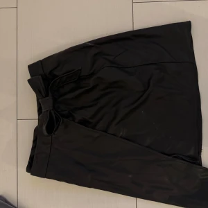 Svart omlottkjol från NA-KD med rosett - Snygg svart omlottkjol från NA-KD i storlek 34. Kjolen har en stilren look med en dekorativ rosett i midjan och är tillverkad i ett material som liknar skinn. Perfekt för dig som vill ha en trendig och edgy vibe.