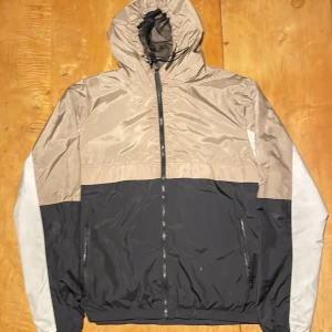 Beige och svart vindjacka med huva - Snygg vindjacka i beige, svart och grått från Fearless Contribution. Jackan har huva med snörning, hel dragkedja framtill och två fickor med dragkedja. Lätt och smidig med meshfoder i huvan, perfekt för blåsiga dagar. Ett litet slitage på justerings snöret där men går säkert att lösa! 