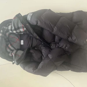 Svart dunjacka från Burberry - Snygg svart dunjacka från Burberry med klassiskt rutigt foder i grått, vitt och rött. Jackan har huva, dragkedja och tryckknappar framtill samt en puffig, quiltad design som håller dig varm. Perfekt för kalla dagar och riktigt stilren look. Man kan även ta av armarna så det blir till en väst. Kvitto finns!