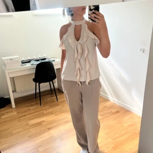 Beige volangtop med halterneck - Supersnygg beige halterneck-top från Missay med volangdetaljer framtill och öppen rygg. Toppen är ärmlös och har en mjuk, flowy känsla i lätt polyester. Perfekt för dig som gillar en chic och trendig look med extra detaljer. Passar perfekt tilll krök eller andra uppklädda sammanhang! 