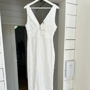 Vit långklänning med blommigt mönster från Adoore - Supersnygg vit långklänning från Adoore med v-ringad hals och ärmlös design. Klänningen har ett diskret blommigt mönster i broderad stil och är tillverkad i bomull. Perfekt för sommarens alla tillfällen och ger en fräsch och romantisk vibe.