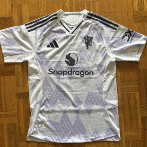 Manchester United Cunha 10 Fotbollströja - Helt ny snygg Manchester United bortatröja med Cunha 10 på ryggen. Tröjan är vit med ljuslila geometriskt mönster, svarta Adidas-ränder på axlarna och Snapdragon-logga på bröstet. Tillverkad i lätt och ventilerande polyester, perfekt för match eller träning. storlek XL men passar L