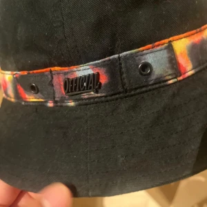 Svart bucket hat från Official - Bucket hat i svart bomull med färgglad batikmönstrad detalj runtom och metallmärke med 'Official'. Snygg och chill stil som passar perfekt till streetwear. Hatten har rund form och ventilerande öljetter på sidorna.