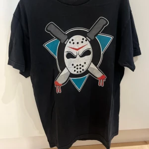 Svart t-shirt med mask och knivar - Cool svart t-shirt med tryck av en hockeymask och två korsade knivar med bloddetaljer. Grafiskt motiv i vitt, rött och blått på bröstet. Klassisk rund hals och korta ärmar, perfekt för dig som gillar edgy streetstyle.