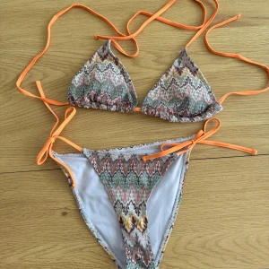Mönstrad bikini med orange snören - Snygg bikini med färgglatt mönster i rosa, grön, gul och grå toner. Triangeltopp och trosa med knyt i sidorna och tunna orange snören. Perfekt för stranden eller poolen, ger en fräsch och trendig look.