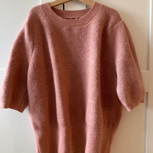 Rosa stickad tröja med halvärm - Mysig stickad tröja i en dämpad rosa nyans. Tröjan har rund halsringning, breda ribbade muddar och halvärmar som ger en avslappnad vibe. Perfekt för lager-på-lager och chill dagar. Materialet är mjukt och känns skönt mot huden.