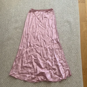 Ljusrosa långkjol i satin - Supersnygg ljusrosa långkjol i glansigt satinmaterial. Kjolen har en enkel, flowy siluett och ett elastiskt midjeband med spetsdetalj upptill. Perfekt för dig som gillar en feminin och trendig look. (Den är väldigt skrynklig på denna bilden)💗🌸