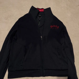 Svart zipjacka från Hugo Boss - Svart zipjacka från Hugo Boss med röd logga på bröstet och röda detaljer på insidan. Jackan har hög krage, hel dragkedja och två fickor. Tillverkad i mjuk bomullsmix, perfekt för en clean och sportig look.