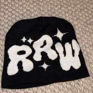 Svart mössa med vit RRW-tryck - Cool svart mössa med stora vita bokstäver 'RRW' och stjärndetaljer framtill. Stickad i mjukt material och perfekt för att ge din outfit en streetstyle-vibe.