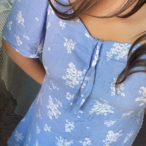 Blå blommig klänning - Supersöt ljusblå klänning från lager med vita blommor, korta ärmar och knyt framtill vid halsen. Klänningen har en  v-ringad urringning och är gjord i mjuk bomull, superluftig och bra för varma dagar. Passformen är normal och längden är kort.