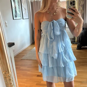 Ljusblå axelbandslös volangklänning - Superfin ljusblå klänning från Nelly med flera lager volanger och axelbandslös design. Framtill har den en stor knytrosett och smock i ryggen för skön passform. Perfekt för sommarens alla tillfällen, lätt och luftig i materialet.🩵🩵