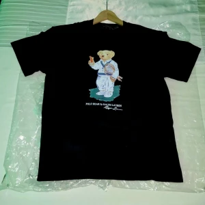 Svart Polo Bear t-shirt Ralph Lauren - Svart t-shirt från Polo Ralph Lauren med ikoniskt Polo Bear-tryck på bröstet där björnen håller en tennisracket och dryck. Klassisk rund hals och korta ärmar. Materialet är mjuk bomull, perfekt för en chill och stilren look.