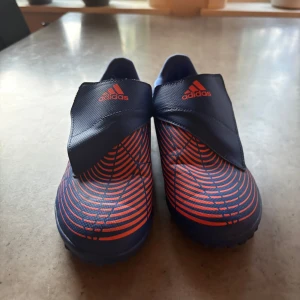 Adidas preditor (röd/orange och blå) - Snygga fotbollsskor från Adidas i mörkblått med röda detaljer och grafiskt mönster. Skorna har en låg profil och täckt snörning för en clean look. Yttermaterialet är syntet och sulan är anpassad för gräs eller konstgräs. Perfekta för dig som vill sticka ut på planen.