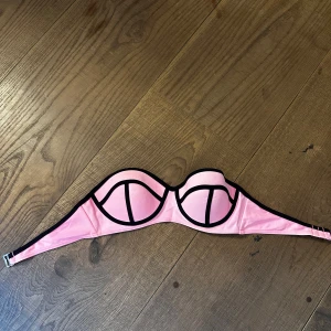 Rosa triangl liknande bikinitopp - Snygg rosa bikiniöverdel med svarta kontrastkanter och coola grafiska linjer över kuporna. Bandeau-modell med vadderade kupor och justerbar knäppning bak. Perfekt för stranden eller poolen och ger en trendig look.