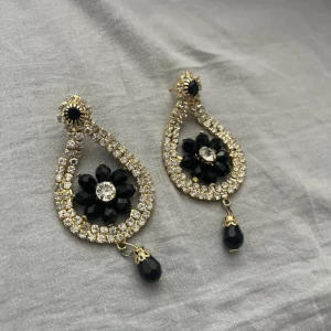 Glittriga örhängen med svart blomma - Säljer ett par droppformade örhängen med glittrande stenar och guldfärgade detaljer. I mitten sitter en svart blomma med en klar sten i centrum och längst ner hänger en svart pärla. Perfekta för att ge din look extra bling och wow-känsla.