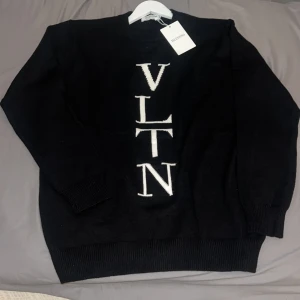 Svart stickad tröja från Valentino - Svart stickad tröja från Valentino med vit VLTN-logga vertikalt framtill. Klassisk rund halsringning och ribbade muddar vid ärmslut och nederkant. Perfekt för dig som gillar stilrena designer och exklusiva märken.