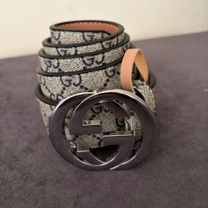 Gucci GG Supreme bälte med silverdetalj - Snyggt Gucci-bälte med klassiskt GG Supreme-mönster i beige och mörkblått. Bältet har en stor, rund spänne i silverfärgad metall med dubbla G-loggan. Tillverkat i canvas med skinn på insidan. Perfekt accessoar för att lyfta din outfit.