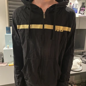 Svart hoodiejacka från BOSS med guld - Svart hoodiejacka från BOSS med dragkedja framtill och huva med snörning. Snygg guldfärgad rand och BOSS-logga över bröstet. Tillverkad i mjuk bomull, perfekt för en chill och stilren look. Storlek S.