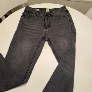 True Religion grå jeans Joey Super T - Säljer ett par grå jeans från True Religion, modell Joey Super T. De har markerade sömmar, snygga bakfickor med lock och logga, samt klassisk femficksdesign. Jeansen är i tvättad grå nyans och har raka ben. Perfekta för dig som gillar streetwear vibes.