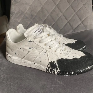 Maison Margiela sneakers med färgstänk - Unika Maison Margiela sneakers i vitt canvasmaterial med svarta färgstänk och råa kanter. Skorna har snörning, rund tå och coola detaljer på sulan. Märkets ikoniska etikett syns på plösen. Perfekta för dig som gillar statement pieces och streetstyle.