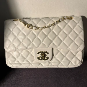 Vit quiltad Chanel handväska i skinn - Supersnygg vit handväska från Chanel med klassiskt quiltat mönster och guldfärgad kedja. Väskan har den ikoniska CC-loggan på låset och är tillverkad i mjukt skinn. Perfekt storlek för det viktigaste och har en elegant vibe med exklusiva detaljer.
