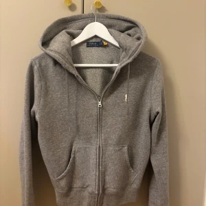 Grå hoodie från Polo Ralph Lauren - Säljer en grå hoodie från Polo Ralph Lauren i storlek S. Tröjan har huva med snörning, dragkedja framtill och två fickor. Klassisk broderad logga på bröstet. Mjuk. Passformen är normal och ärmarna är långa. Nypris: 1999