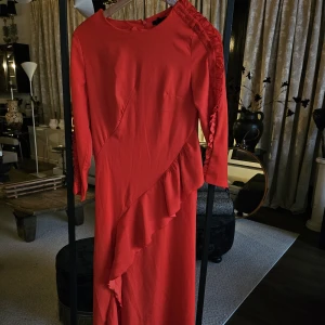 Röd långklänning med volang och knappar - Snygg röd långklänning med volangdetalj som går diagonalt över framsidan. Klänningen har långa ärmar och dekorativa knappar längs ena axeln och ärmen. Materialet känns mjukt och lätt, perfekt för att sticka ut med en färgstark look.