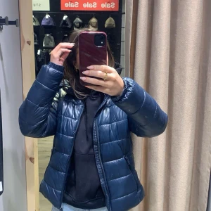 Mörkblå pufferjacka med dragkedja - Säljer en mörkblå pufferjacka med glansig finish och quiltad design. Jackan har lång ärm, hög krage och dragkedja framtill. Perfekt för kyliga dagar och lätt att matcha med olika outfits.