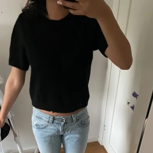 Svart croppad stickad tröja - Svart, croppad stickad tröja med korta ärmar. Enkel och stilren design som passar perfekt till jeans eller kjol. Mjuk och skön att ha på sig, perfekt för dig som gillar en clean och avslappnad look.