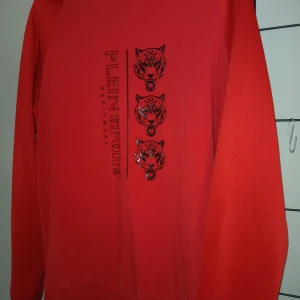 Röd sweatshirt från Plein Sport XL - Snygg röd sweatshirt från Plein Sport i storlek XL. Tröjan har tryck med tre svarta tigerhuvuden och logga på framsidan. Långärmad med ribbade muddar och rund hals. Perfekt för dig som gillar streetwear och sportig stil.