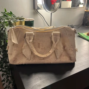 Beige axelväska i jeans med kedja - Säljer denna beigea axelväska i jeansmaterial med slitna detaljer och silverfärgad kedjerem. Väskan har både handtag och axelrem, samt dragkedja upptill. Den är endast använd några fåtal gånger och är därför i nyskick.