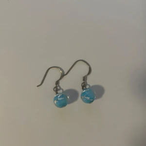 Blå pärlörhängen med silverdetaljer - Säljer ett par snygga örhängen med blå runda pärlor och silvriga inslag. Krokarna är i metall och pärlorna har ett unikt mönster med vita och silverfärgade slingor. Perfekt för dig som vill ha något färgglatt och personligt i öronen.