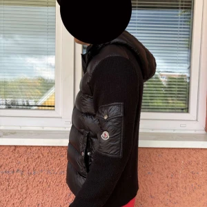 Svart dunväst med huva från Moncler - Säljer en svart dunväst från Moncler med stickade ärmar och huva. Västen har quiltad framsida i glansigt material, dragkedja och en ficka med Moncler-logga på ärmen. Perfekt för lager-på-lager och snygg streetstil.