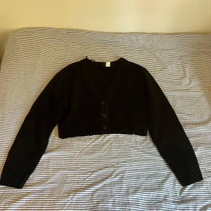 Svart kofta från H&M  - Svart croppad kofta från H&M med v-ringning och tre knappar framtill. Stickad i mjukt material med ribbade muddar vid ärmslut och nederkant. Aldrig använd.