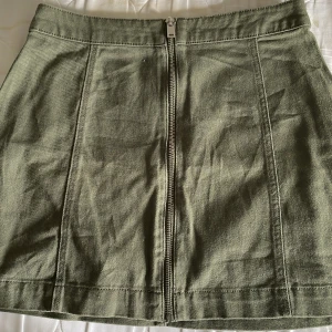 Olivgrön kortkjol från H&M Divided - Snygg olivgrön kortkjol från H&M Divided i storlek 36. Kjolen har en rak passform och är tillverkad i ett slitstarkt bomullsmaterial. Framtill finns en synlig dragkedja som ger en cool detalj. Perfekt att matcha med sneakers eller boots.