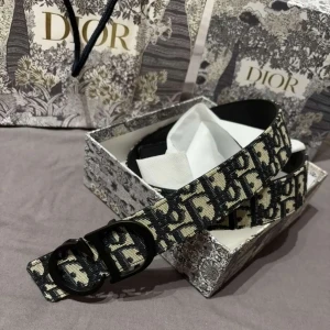 Svart och beige Dior bälte med logomönster - (Möts upp i stockholm). Bälte från Dior med ikoniskt svart och beige logomönster. Bältet har en bred design och en svart metallspänne formad som ett D. Materialet är textil med detaljer i skinn på insidan. Perfekt accessoar för att lyfta din outfit.