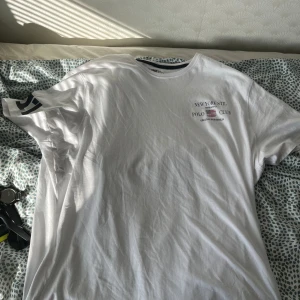 Vit t-shirt från New York Stil Polo Club - Vit t-shirt i bomull från New York Stil Polo Club med tryckt amerikansk flagga och text på bröstet. Klassisk rund halsringning med mörkblå kant och tryckt siffra 17 på ena ärmen. Perfekt för en sportig och avslappnad look.