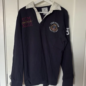 Mörkblå pikétröja från Lexington - Snygg mörkblå långärmad pikétröja från Lexington med vit krage och broderade detaljer på bröstet och ärmen. Klassisk rugbyinspirerad stil med knappar i halsen och coola brodyrer. Perfekt för dig som gillar preppy och sportig look.