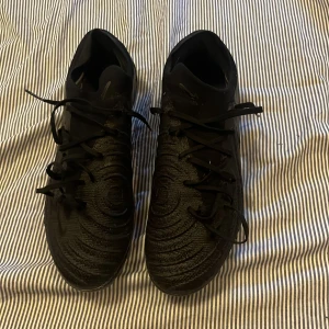 Nike phantom GX ll Elite FG Shadow - Säljer ett par helsvarta Nike fotbollsskor med snygg textur och klassisk swoosh på sidan. Skorna har snörning och mönstrad ovandel i syntetmaterial för bra grepp och känsla på planen. Yttersulan har fasta dobbar för spel på gräs.