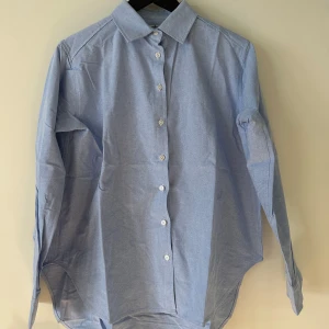 Ljusblå skjorta från Totême - Klassisk ljusblå skjorta från Totême med rak passform och tydlig krage. Skjortan har vita knappar framtill och broderad logga på bröstet. Tillverkad i bomull, perfekt för en clean och stilren look.